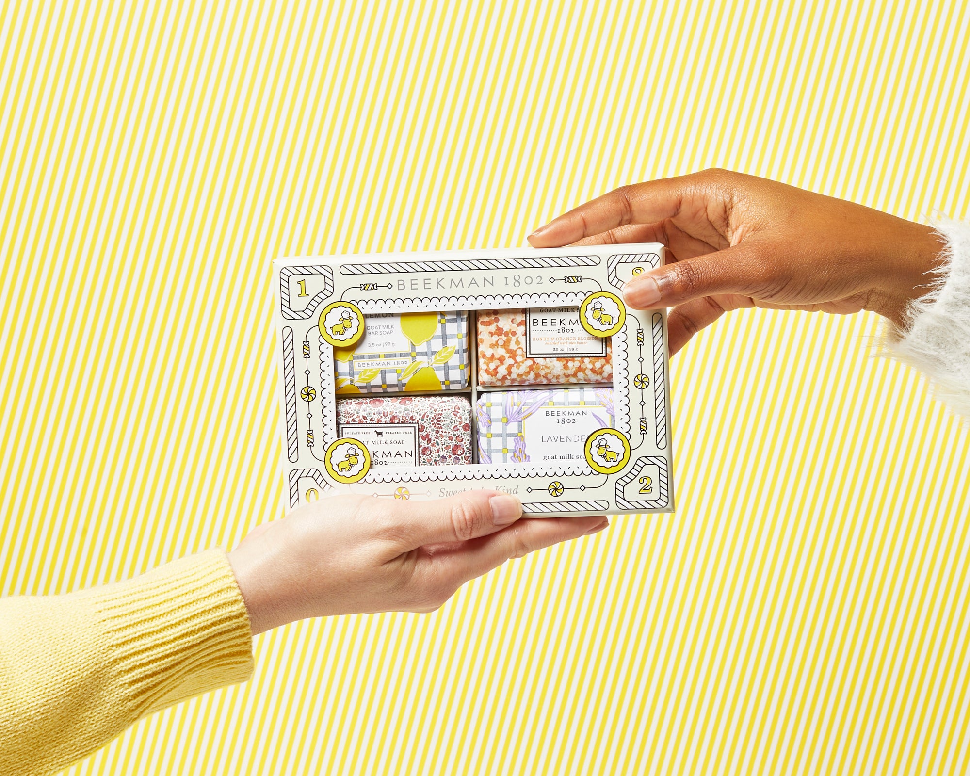 Hand gifting Beekman 1802's Sweet Travels 4-Piece Travel Size Bar Soap Set, which shows sunshine lemon, honey and orange blossom, honeyed grapefruit, and lavender 3.5 ounce travel-sized bar soaps on the front of the box, to another hand, on a yellow and white striped background.