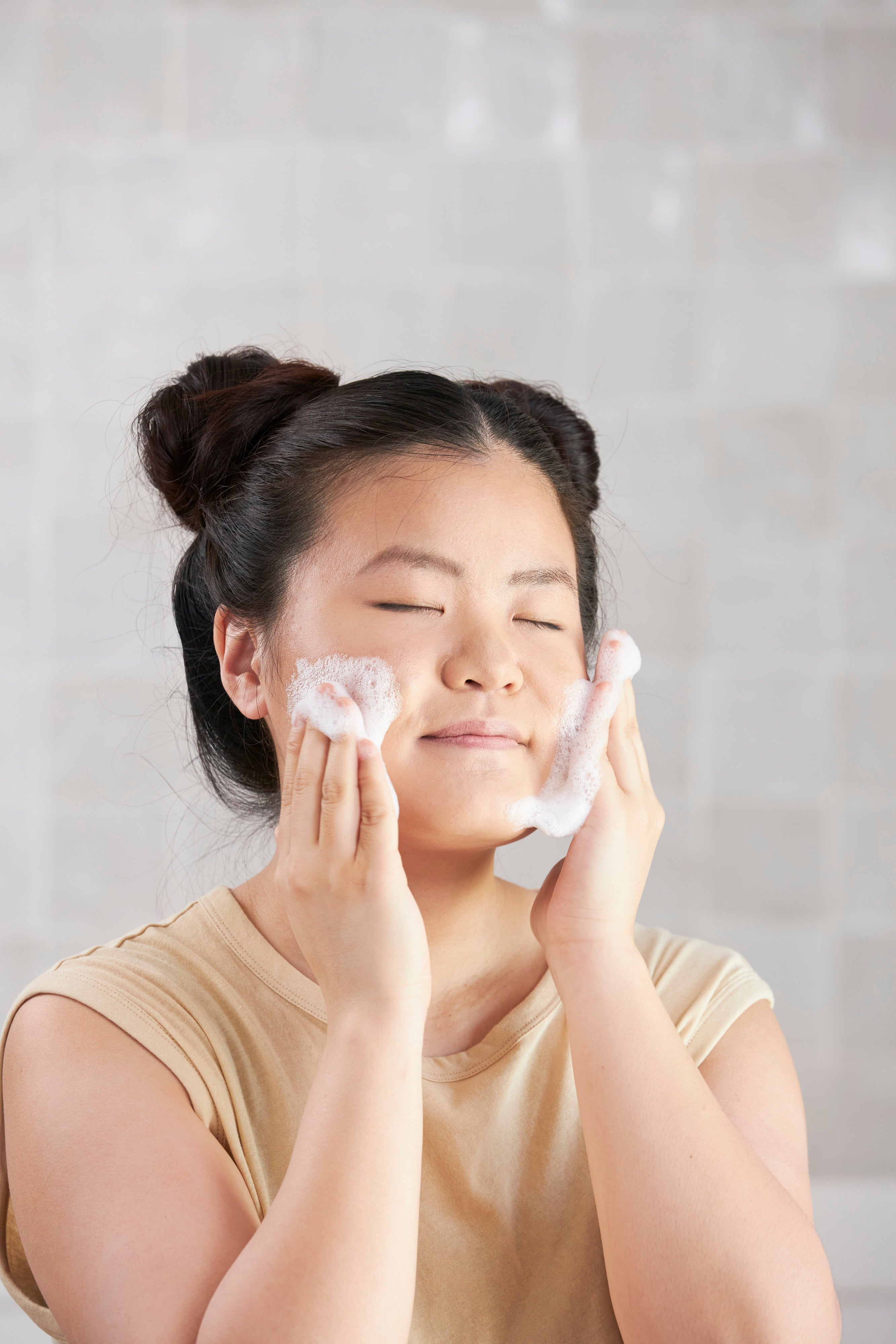 Image of model closing her eyes and applying Beekman 1802's Milk bar facial cleansing bar formula to her face with her hands creating suds, on a grey background.